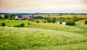 A view of a rural landscape in Northwest Ohio, showing large homes on a grassy hillside with hay bales and a pond in the distance.