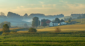 A scenic view of a rural farm in Southeast Ohio, with fields of crops, rolling hills, and houses surrounded by early morning mist under a pale sky.